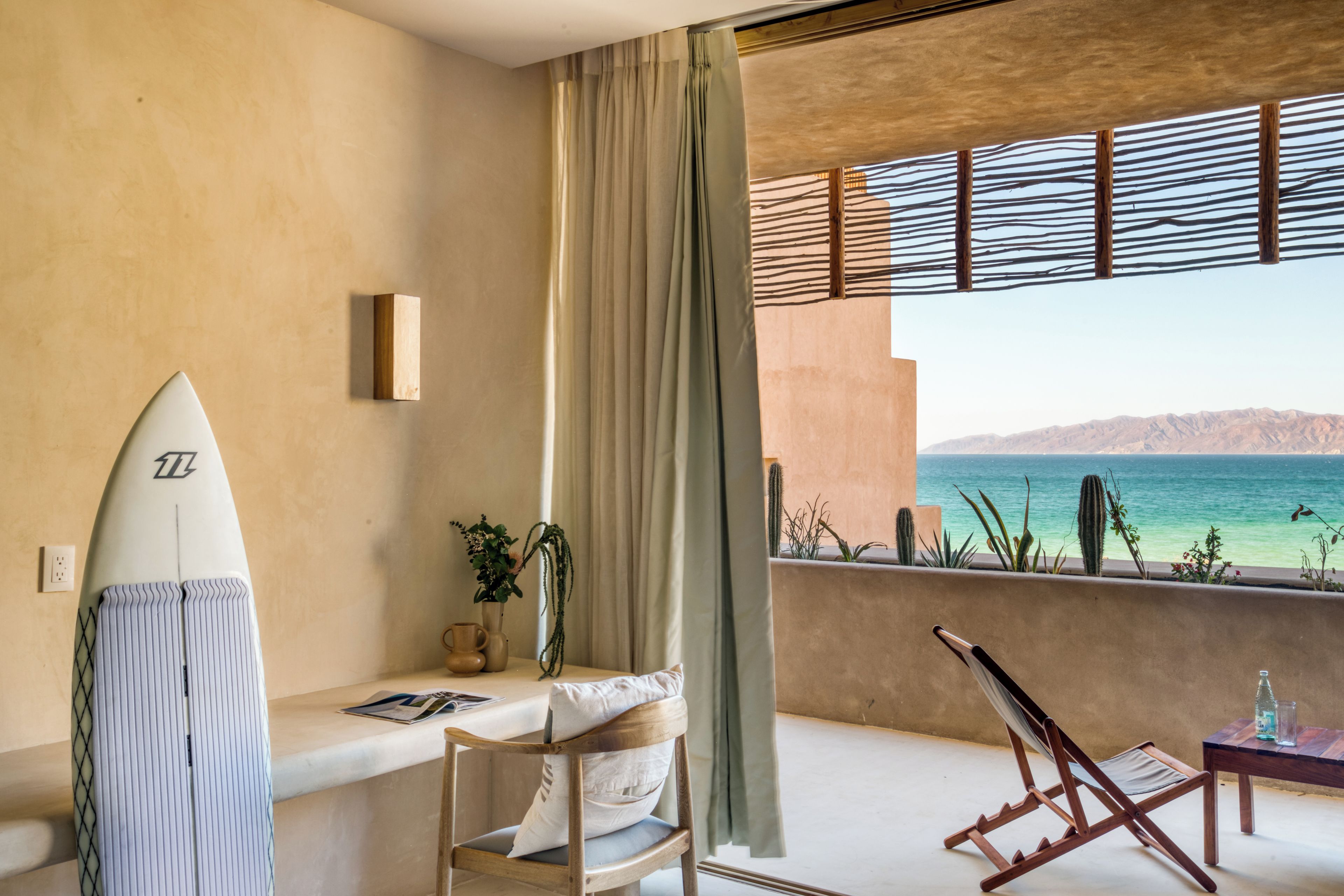 Una habitación con vista al mar, una tabla de surf, una silla, una mesa y un ventanal con vista a cactus.
