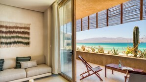 A modern living room with a sofa, a coffee table, and a balcony with a view of a beach and cactus. - Amina Wind Resort (La Ventana)