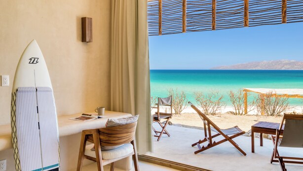 A beachside room with a surfboard, a table with chairs, and a view of the ocean. - Amina Wind Resort (La Ventana)