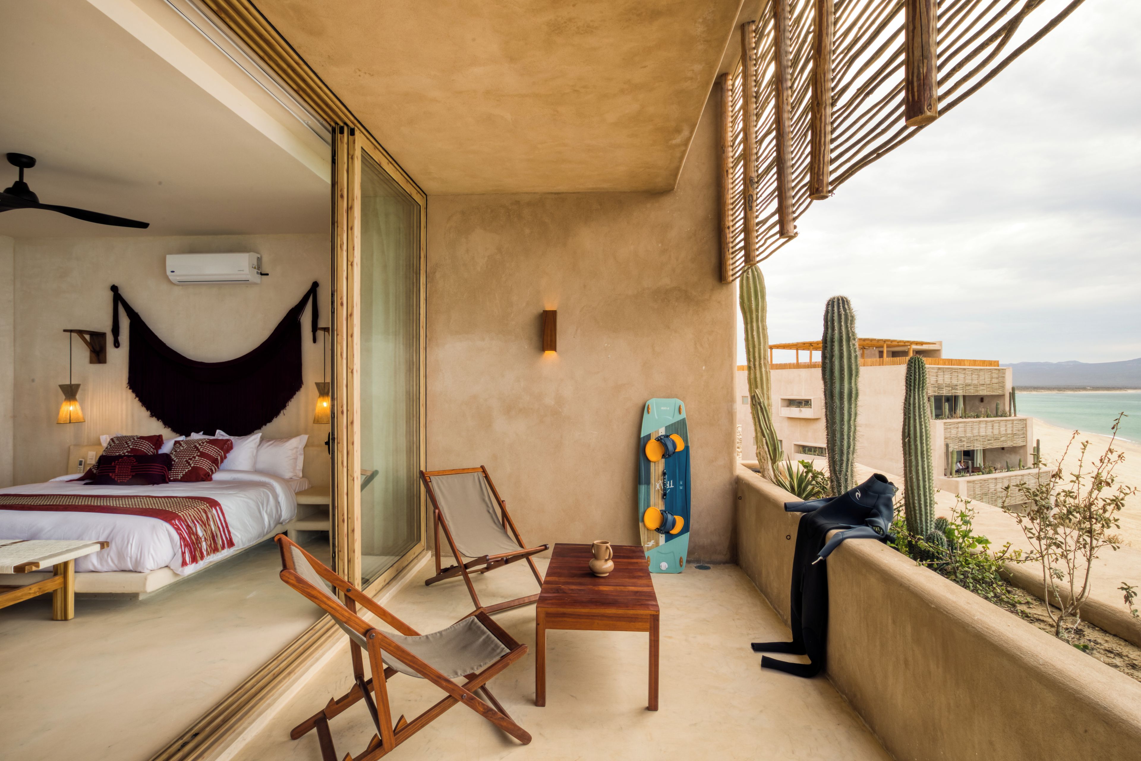 A balcony with a bed, two chairs, a table, a surfboard, and a cactus.