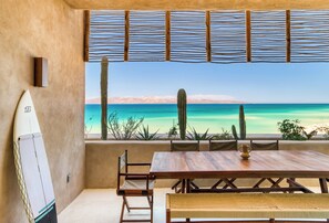 A wooden table with chairs, a surfboard, and a view of the ocean and desert landscape.