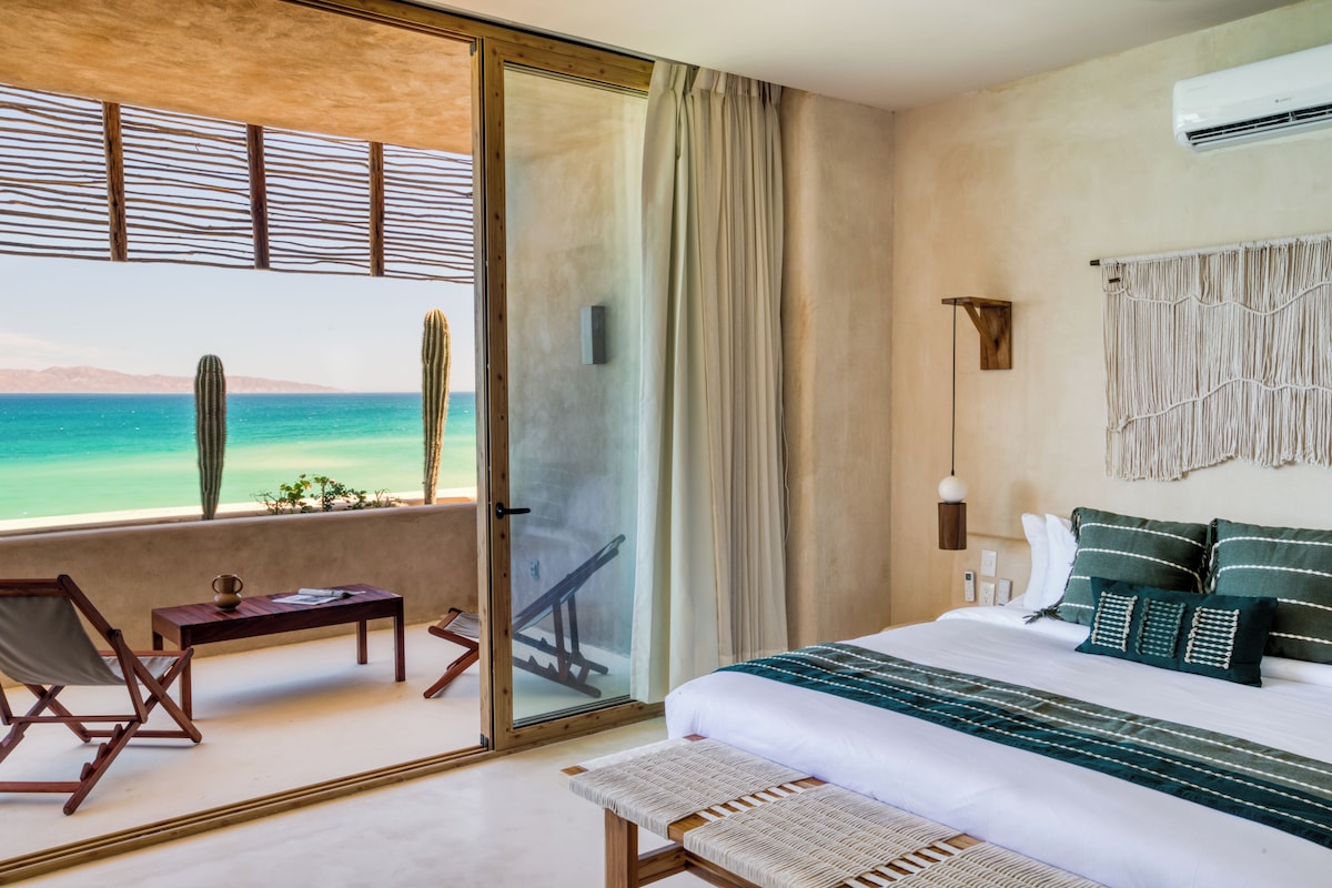 Un dormitorio con cama, un banco, una mesa, una silla, vistas al océano y un balcón.