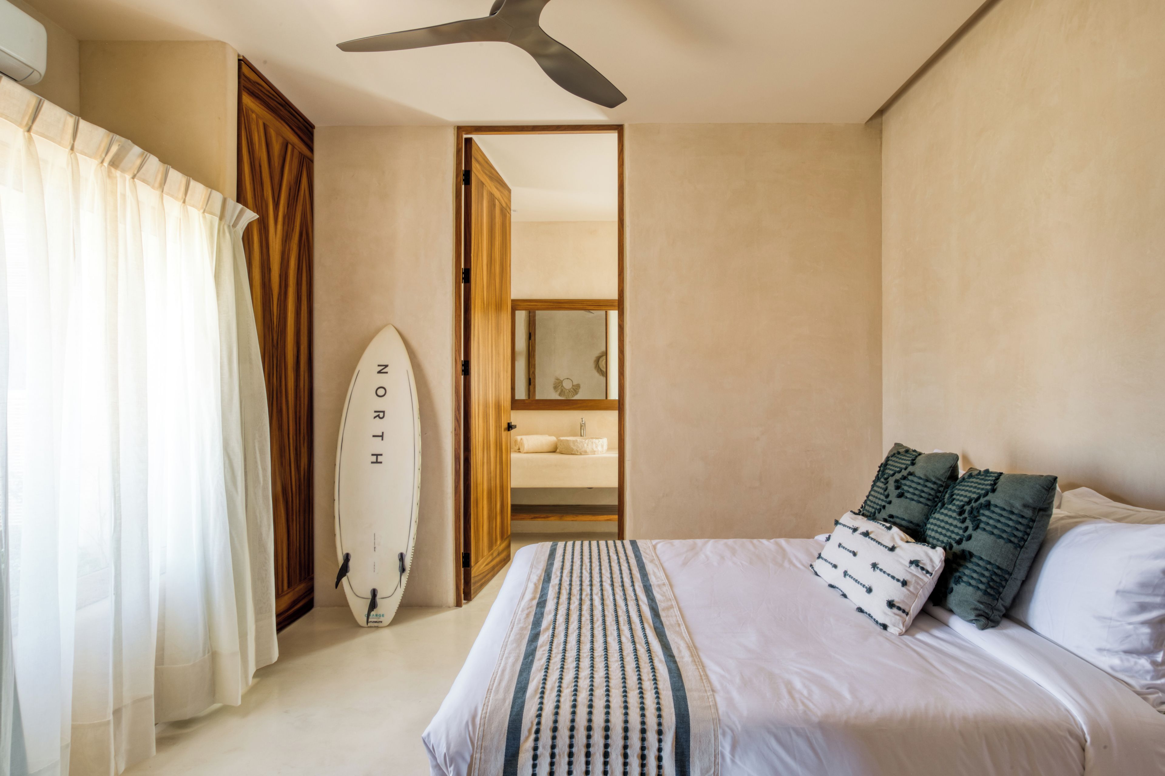 A bedroom with a bed, a ceiling fan, a surfboard, and a bathroom visible through an open door.