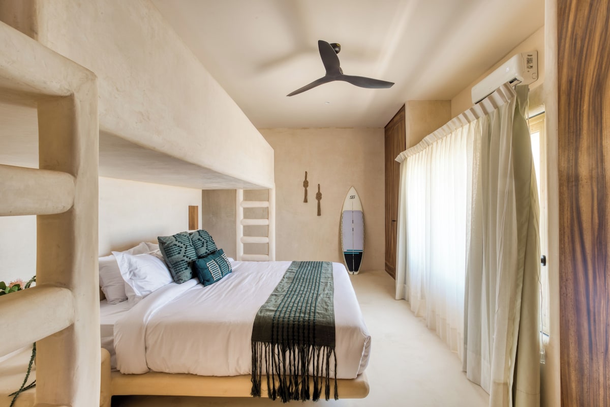 Un dormitorio con cama, almohadas, ventilador de techo, tabla de planchar en la pared y ventana con cortinas.