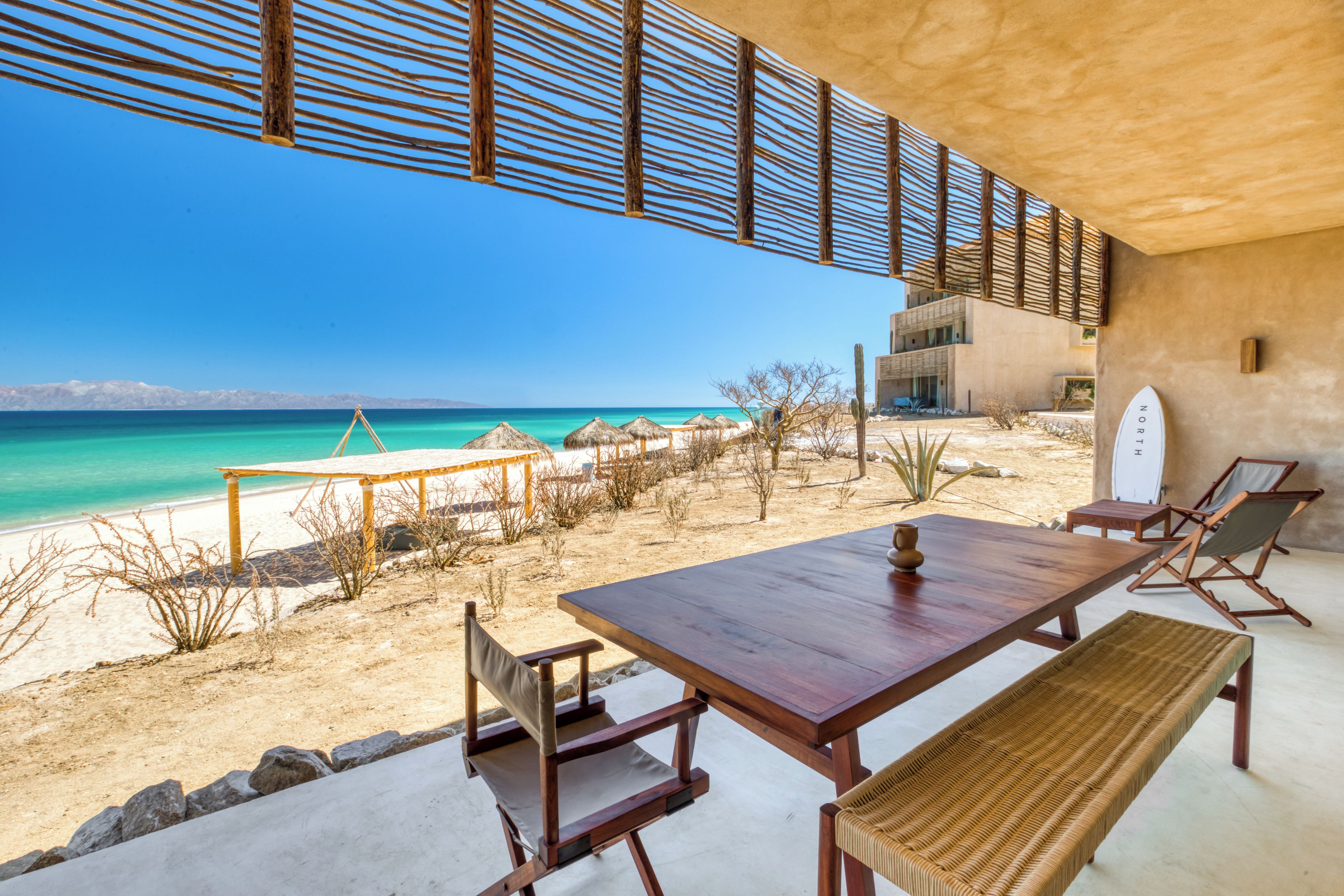 A beachside dining area with a wooden table and chairs, a bench, and a view of the ocean.