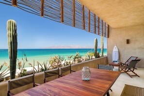 Una mesa de madera con un tarro de vidrio, una tabla de surf y una silla en un balcón con vistas a una playa y un paisaje desértico con cactus.