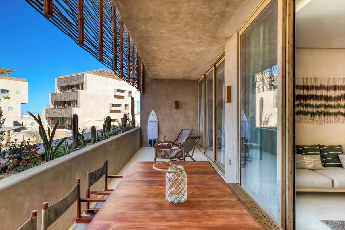 Un balcón con mesa de madera, sillas, una tabla de surf y un muro de vidrio con sofá.