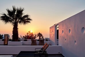 A rooftop terrace with a palm tree, lounge chairs, and a modern building with circular cutouts. - Callia Retreat Suites (Santorini)