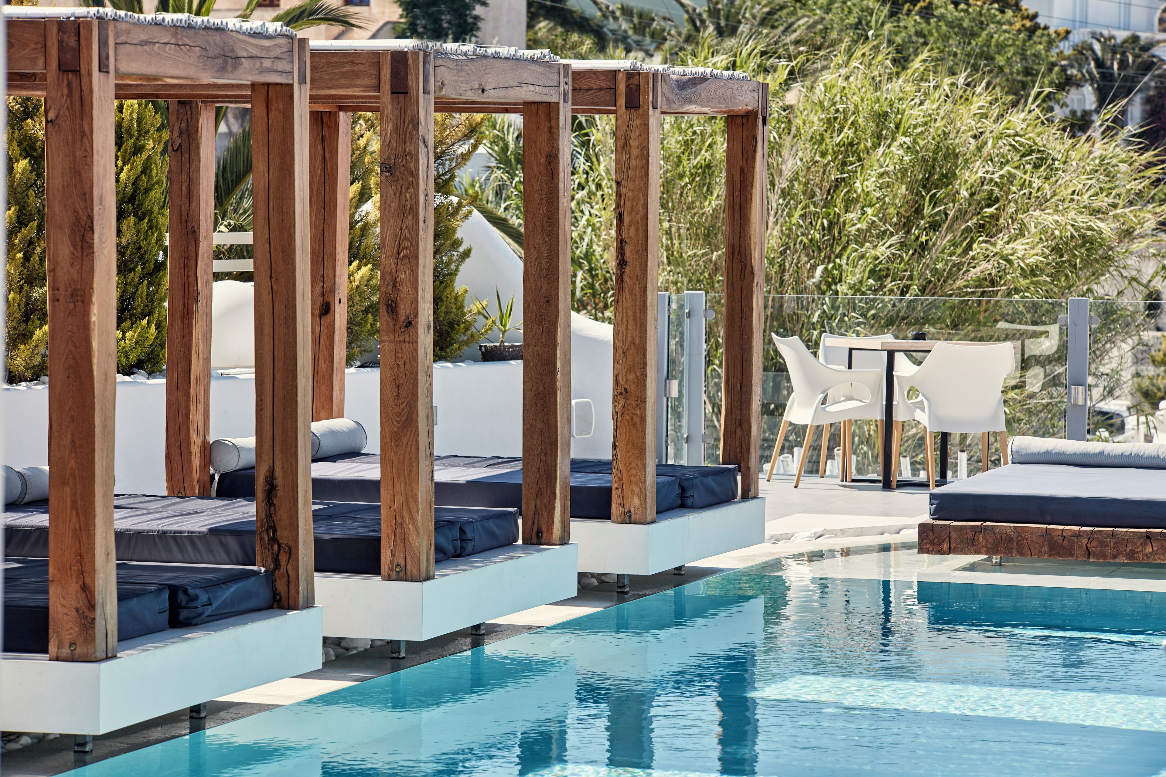 Zona junto a la piscina con pérgolas de madera, sillones de descanso y una pequeña mesa.