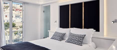 A modern bedroom with a large bed, two patterned pillows, a black headboard, and a view of buildings through a sliding door.