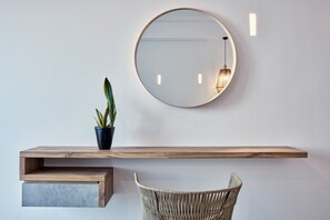 A minimalist room with a wooden console, a round mirror, a potted plant, and a woven chair. - Callia Retreat Suites (Santorini)