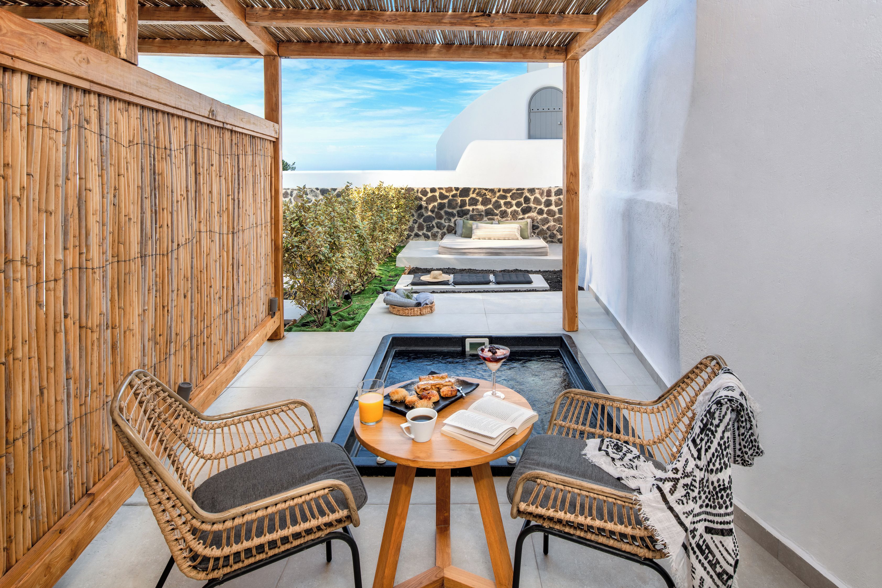 A cozy outdoor seating area with wicker chairs and a table, a small pool, and a view of a white building with a blue sky.