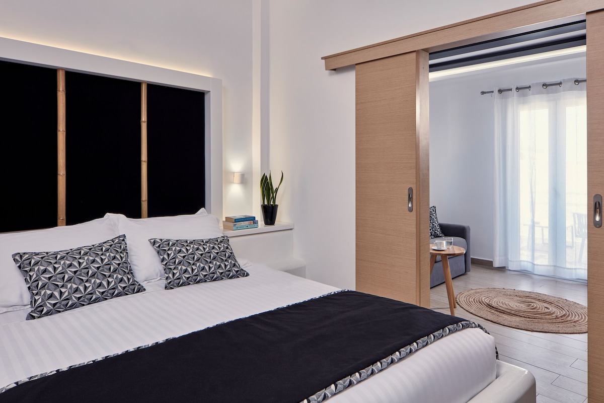 A modern bedroom with a large bed, bedside tables, a potted plant, and a view of a balcony.