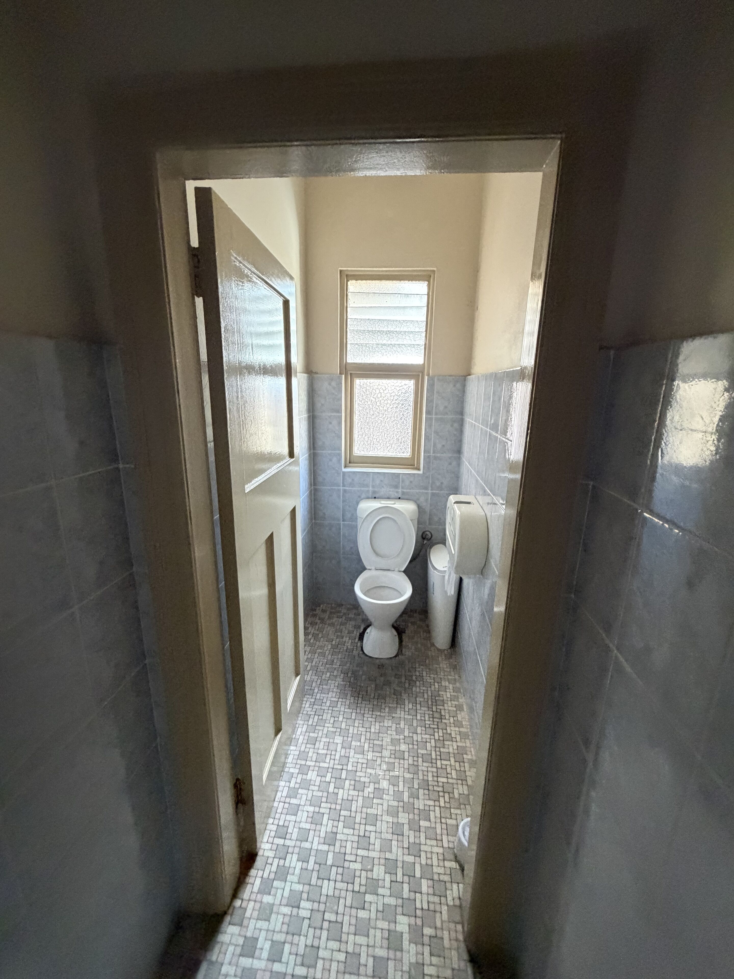 A narrow bathroom with a toilet, a small window, and a tiled floor.