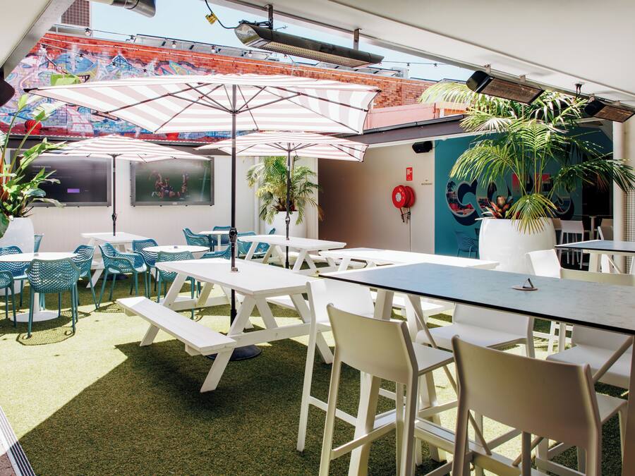 Eine Terrasse im Freien mit weißen Tischen und Stühlen, Sonnenschirmen und Blumentöpfen.