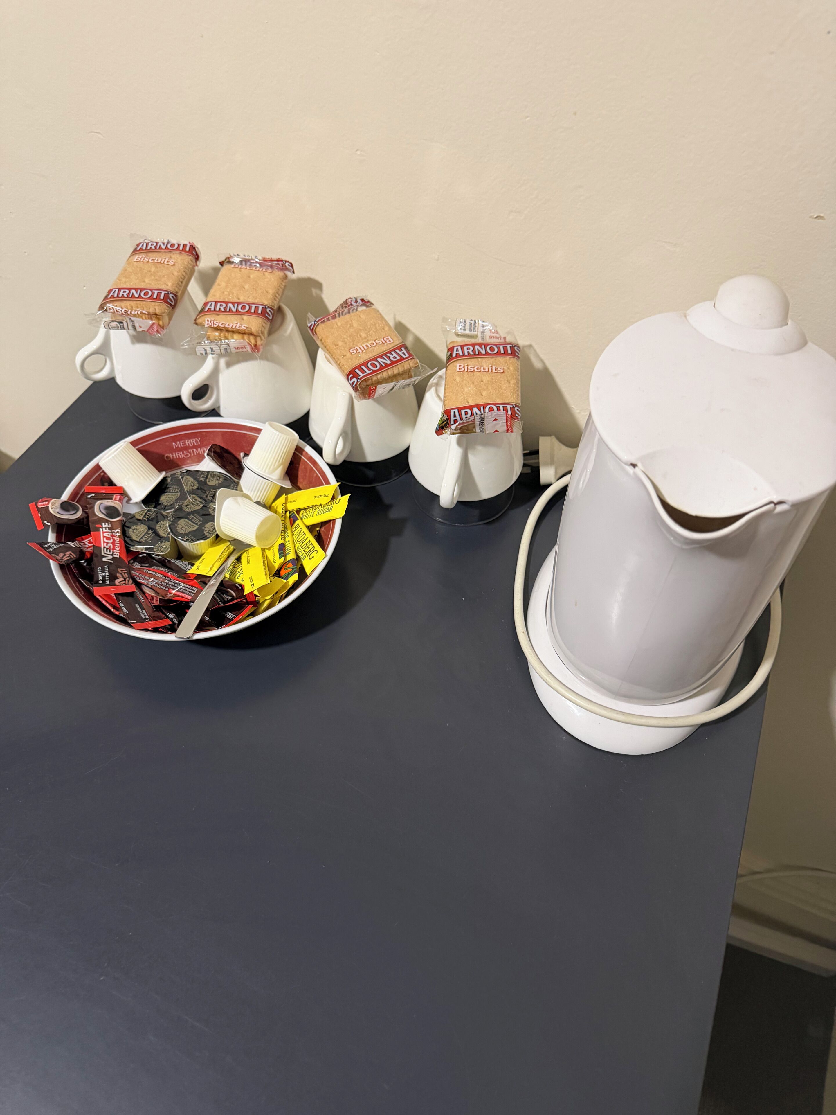 A table with a coffee maker, cups, a bowl of assorted snacks, and biscuit packets.