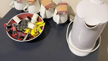 A table with a coffee maker, cups, a bowl of assorted snacks, and biscuit packets.