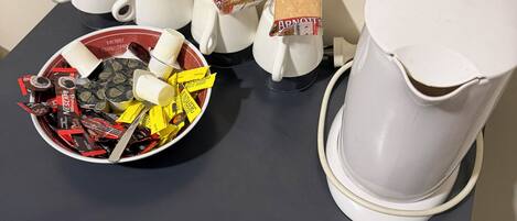 A table with a coffee maker, cups, a bowl of assorted snacks, and biscuit packets.