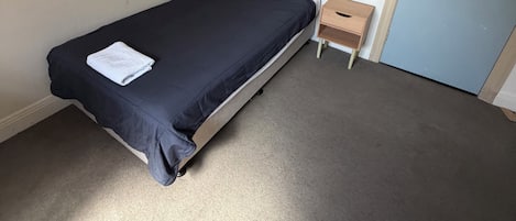 Una habitación pequeña con una cama individual, una cama sencilla, una mesita de noche, un pequeño armario de madera y una puerta con un mapa.