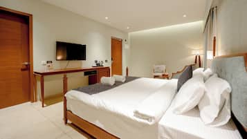 A well-lit bedroom with a large bed, a flat-screen TV mounted on the wall, a desk with a chair, and a door.