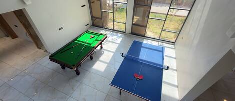 A room with a pool table, a ping pong table, and a window with a view of greenery.