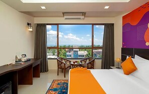 A hotel room with a bed, desk, and chair, a small table with fruit, a window with a view of buildings and palm trees, and an air conditioning unit. - Ira By Orchid Hotels , Porvorim (Alto de Porvorim)