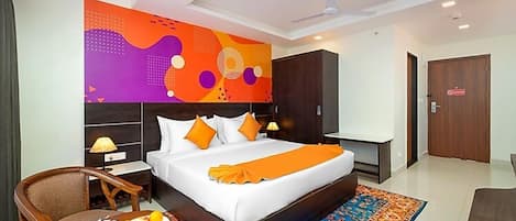 A hotel room with a large bed, a desk, a chair, a small table with fruit, and a colorful rug.