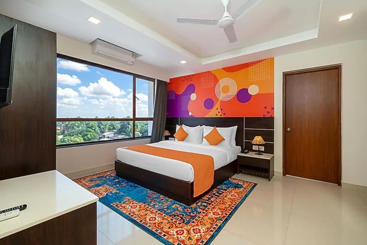 Um quarto com cama, mesa de cabeceira, televisão, ventoinha no teto e uma janela grande.
