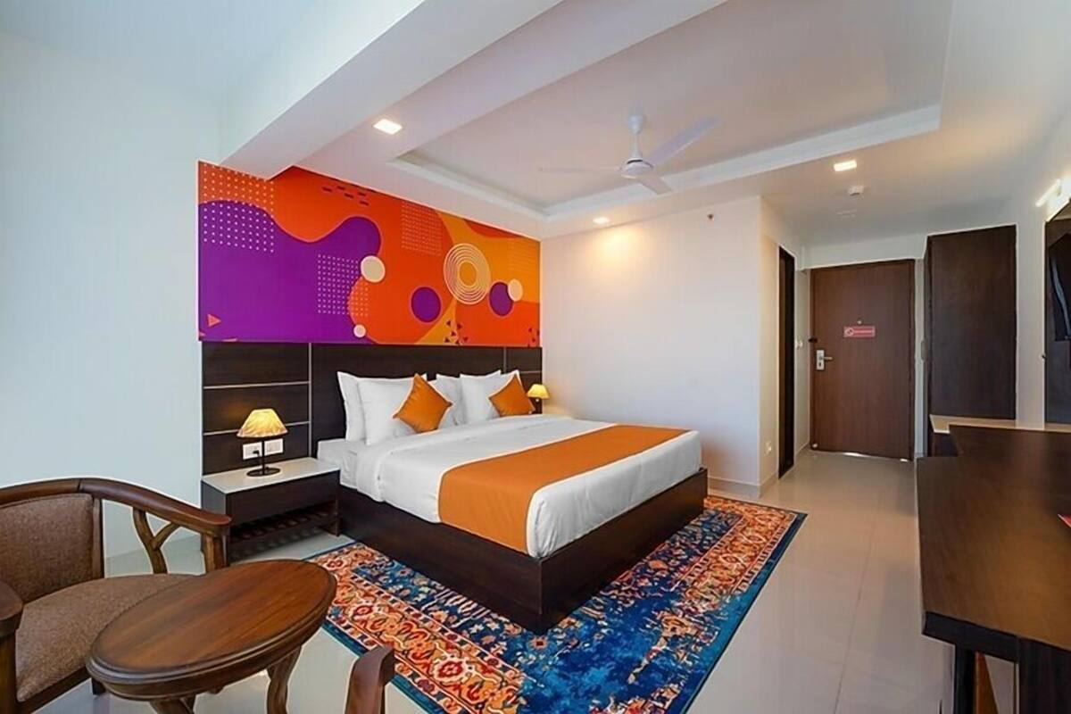 A hotel room with a bed, a desk, a chair, a nightstand, and a wall with a colorful abstract design.