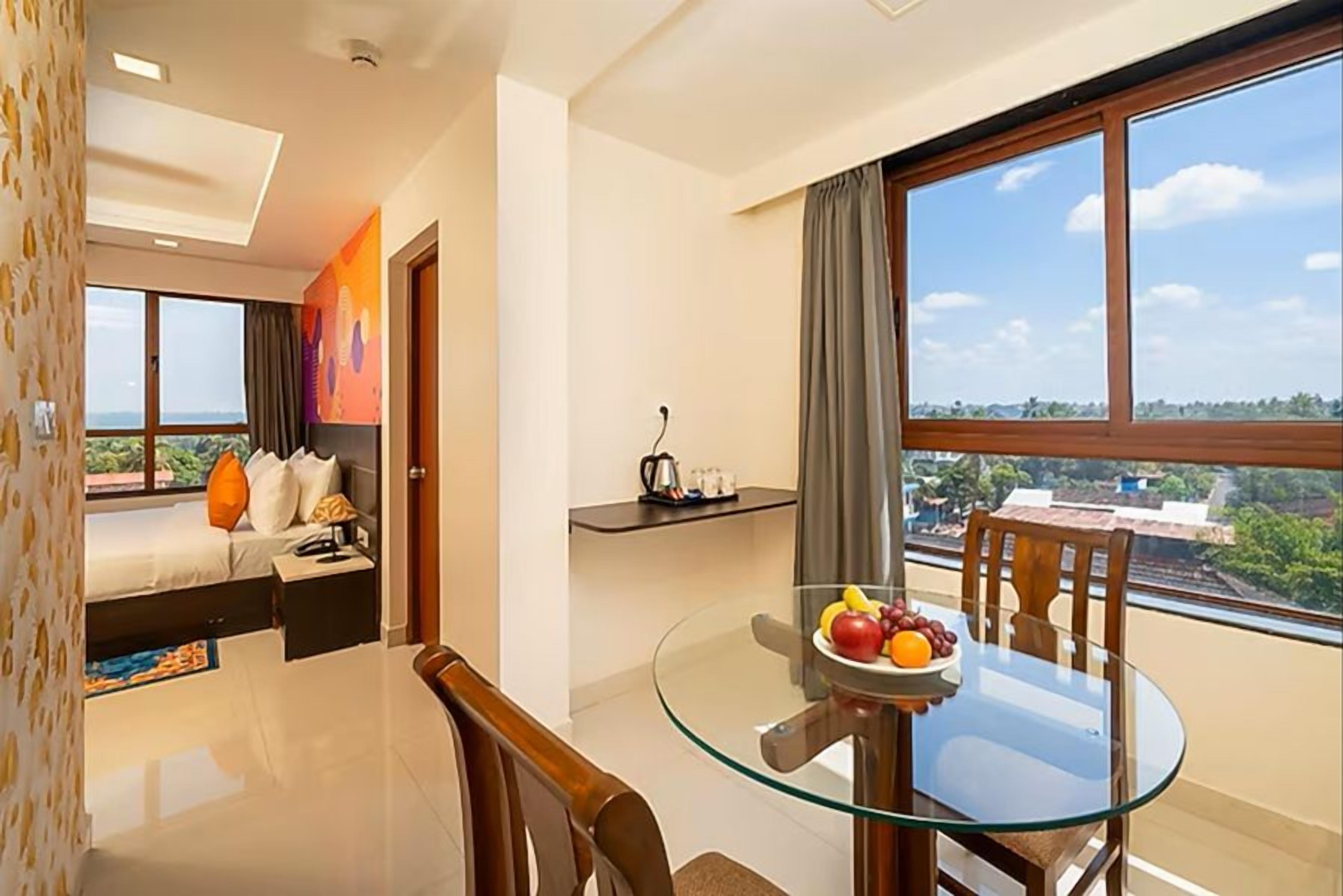 Habitación de hotel con cama, mesa de comedor de cristal con sillas, pequeña cocina y ventana grande con vistas.