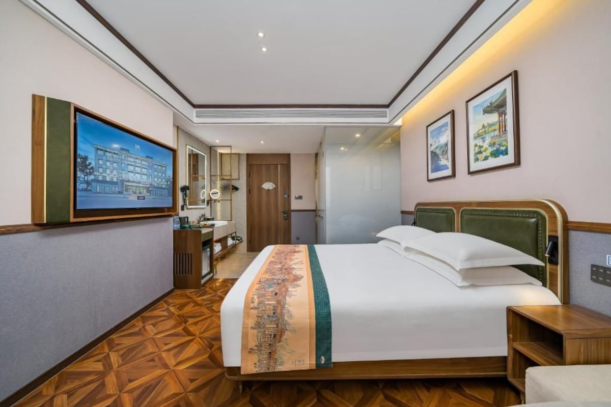 A hotel room with a bed, a desk, a chair, a mirror, and a wall-mounted TV.