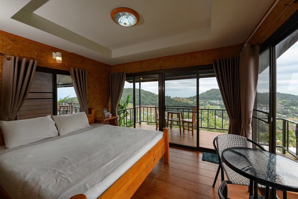 Habitación de hotel con una cama grande, un balcón con mesa y sillas, y vistas a las colinas.