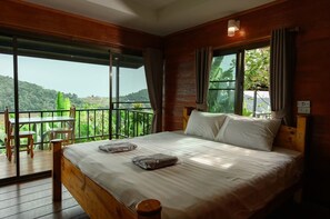 A bedroom with a large bed, a wooden headboard, a balcony with a table and chairs, and a view of greenery. - Mon View Ngarm (Chiang Mai)