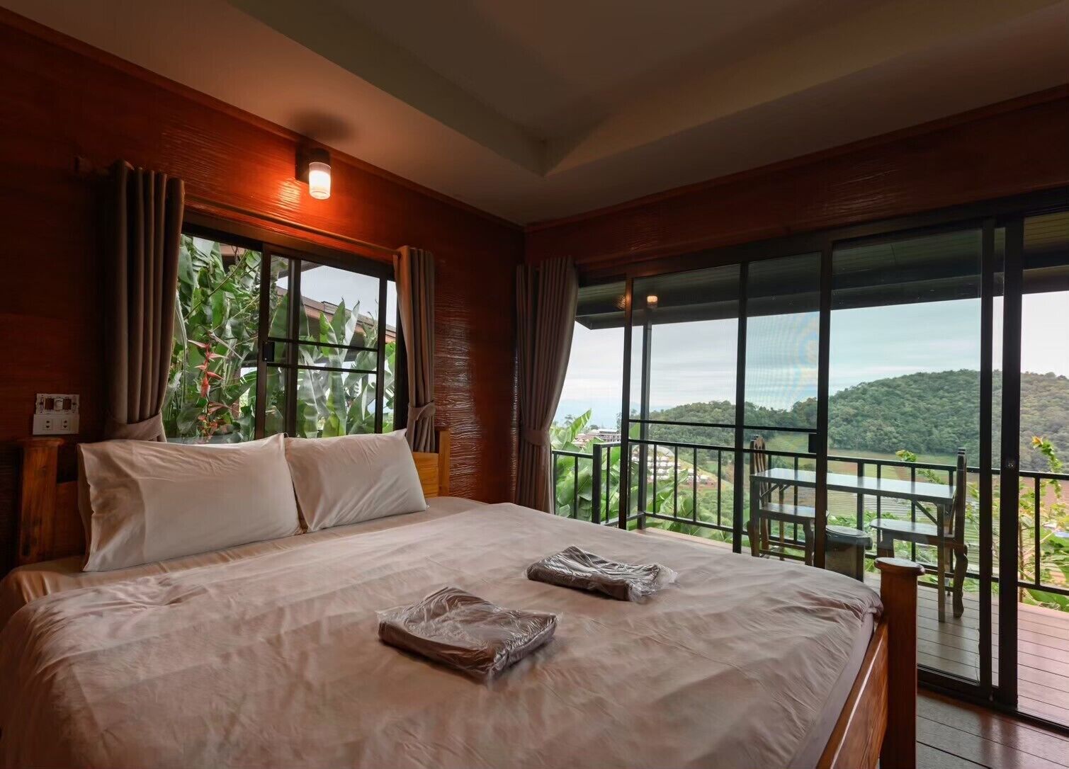 Una cama grande con sábanas blancas, vistas a un paisaje exuberante a través de amplias ventanas y un balcón con mesa y sillas.
