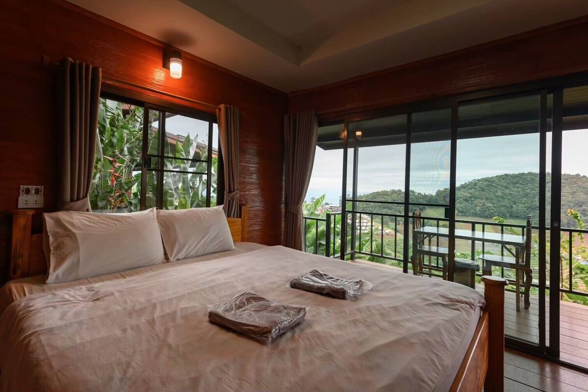 Un grand lit avec du linge de lit blanc, une vue sur un paysage luxuriant grâce à de grandes fenêtres, et un balcon avec une table et des chaises.