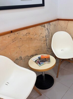 A corner with a white chair, a small round table with magazines, and a marble wall. - Hotel Niza Salou (Salou)