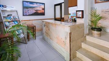 A hotel reception area with a marble counter, a framed picture of a beach, a plant, and a magazine rack.