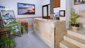 Zona de recepción de hotel con mostrador de mármol, un cuadro enmarco de una playa, una planta y un atril con revistas.