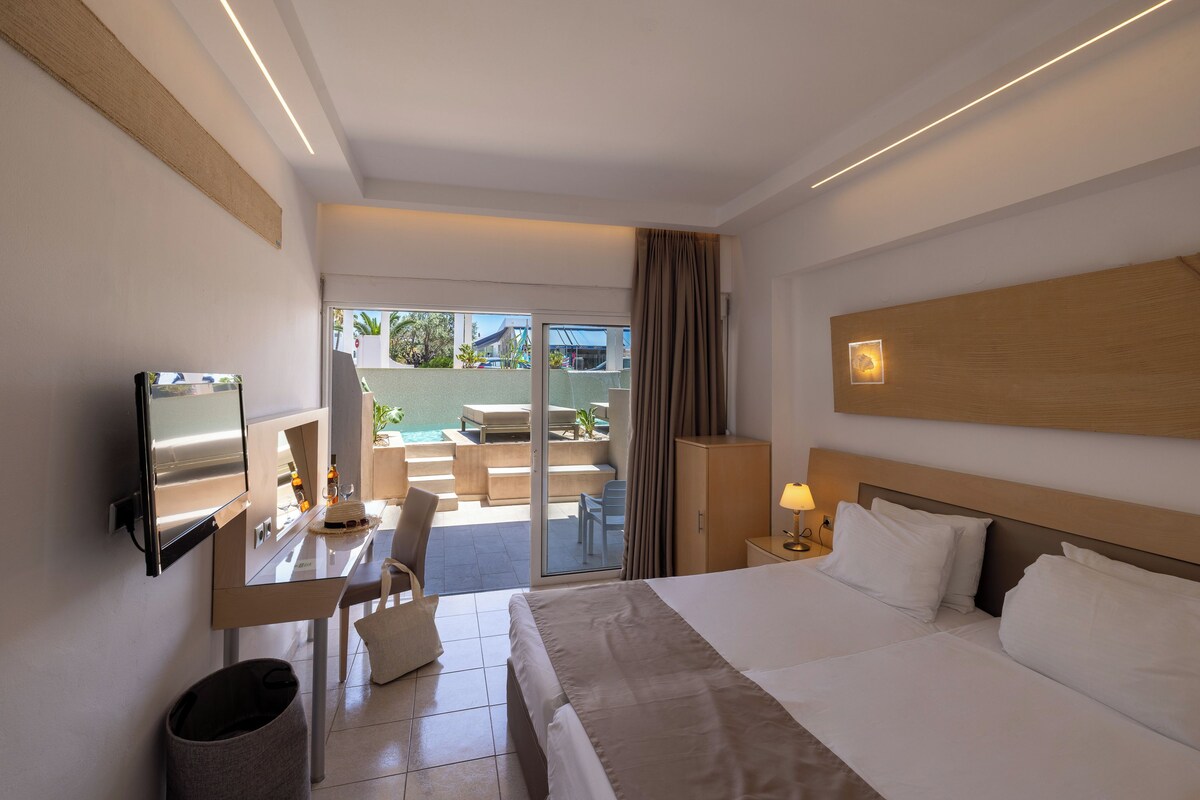 Ein Hotelzimmer mit Bett, Fernseher, Schreibtisch mit Stuhl, Mülleimer und Blick auf einen Außenbereich mit Pool und Sitzgelegenheiten.