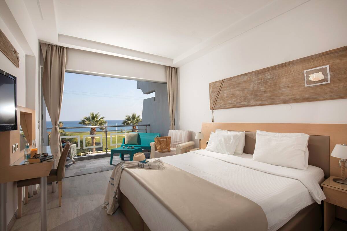 Ein Hotelzimmer mit einem großen Bett, einem Schreibtisch, einem Fernseher und einem Balkon mit Meerblick.