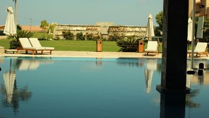 Una piscina con sillones de descanso y sombrillas, un muro de piedra con detalles decorativos, y un cielo despejado.