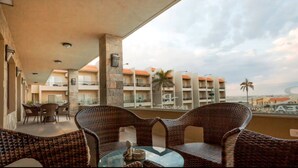 Un balcón con muebles de mimbre y una mesa, con vistas a un edificio con varios balcones y palmeras.