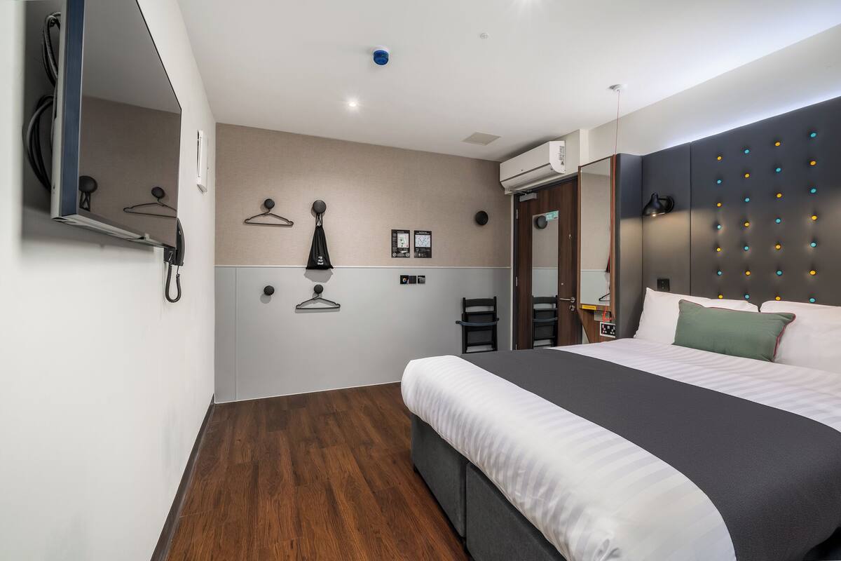 Moderni hotellihuone, jossa on suuri sänky, vaatekaappi, peili ja seinälle asennettu ilmastointilaite.