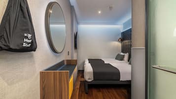 Una habitación de hotel moderna con una cama, una mesita de noche de madera, un espejo, una bolsa negra con la etiqueta "Hot Stuff" y un televisor.