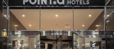 De ingang van een hotel met een glazen gevel, een bord met de tekst 'POINT.A HOTELS' en een lift.
