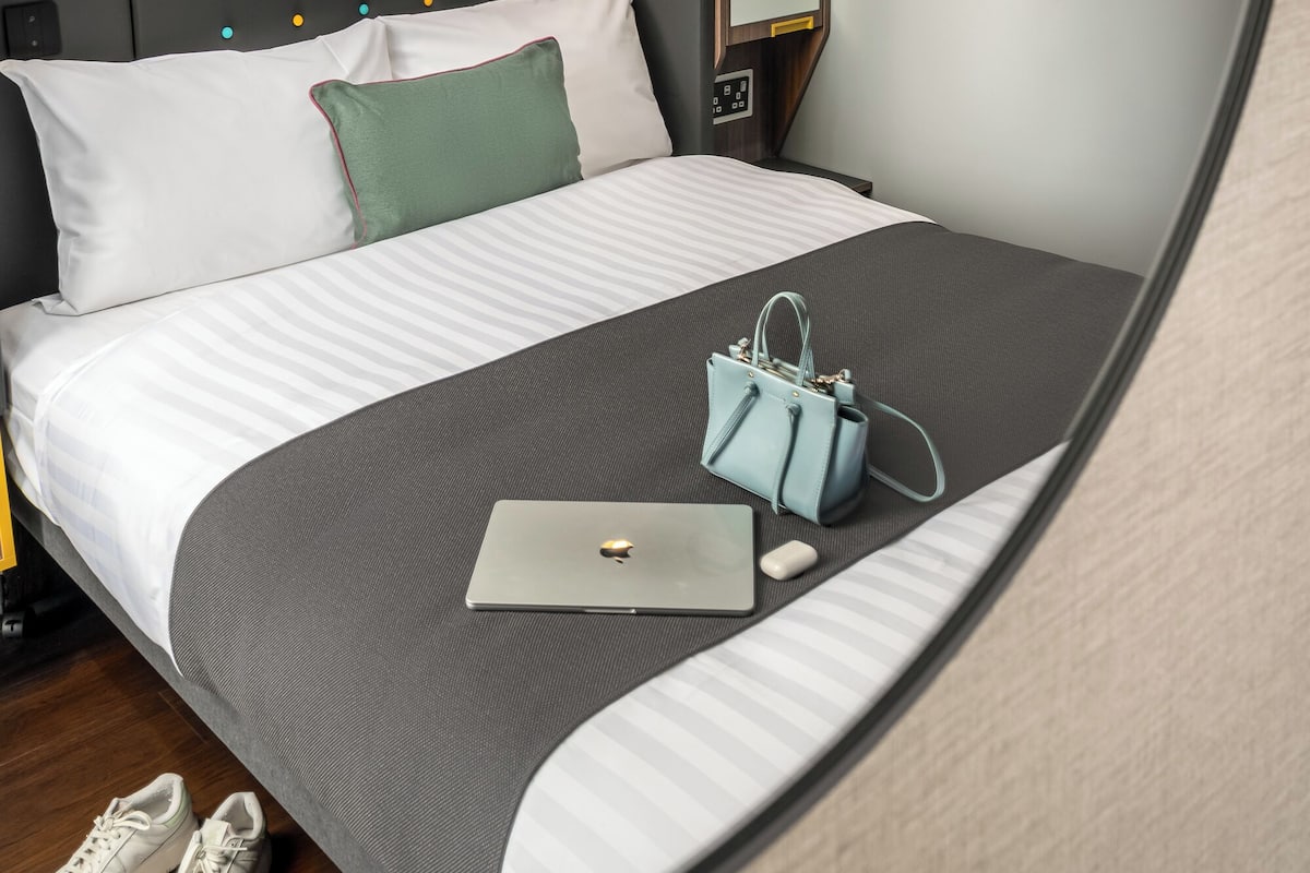 Moderni hotellihuone, jossa on suuri sänky, valaistuilla painikkeilla varustettu pääty, yöpöytä lampun kanssa ja peili.