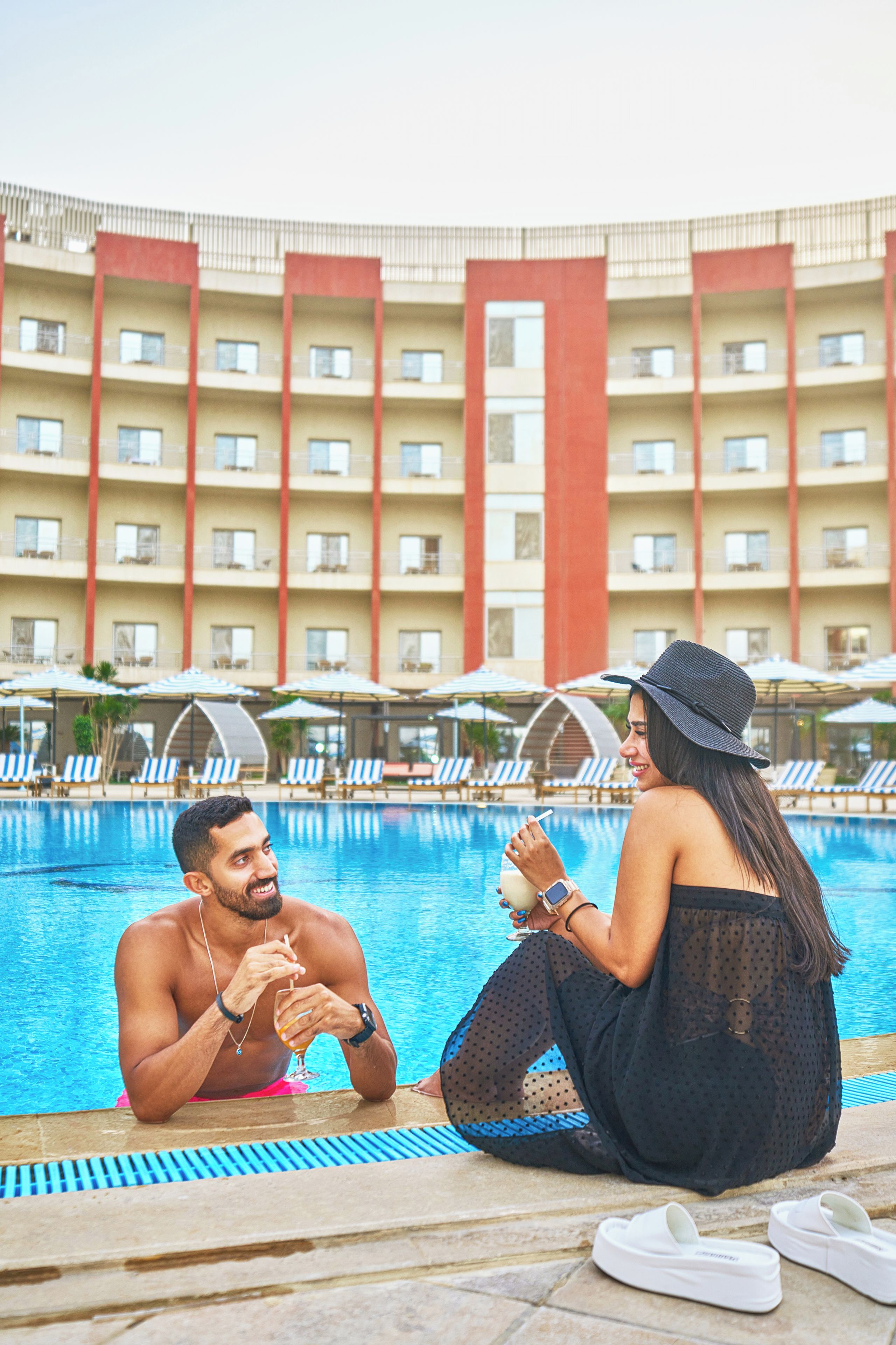 A swimming pool area with lounge chairs and umbrellas, a hotel building with balconies, and a person sitting by the pool.