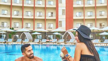 A swimming pool area with lounge chairs and umbrellas, a hotel building with balconies, and a person sitting by the pool.