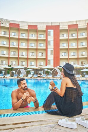 A swimming pool area with lounge chairs and umbrellas, a hotel building with balconies, and a person sitting by the pool. - Tolip El Narges New Cairo (New Cairo)