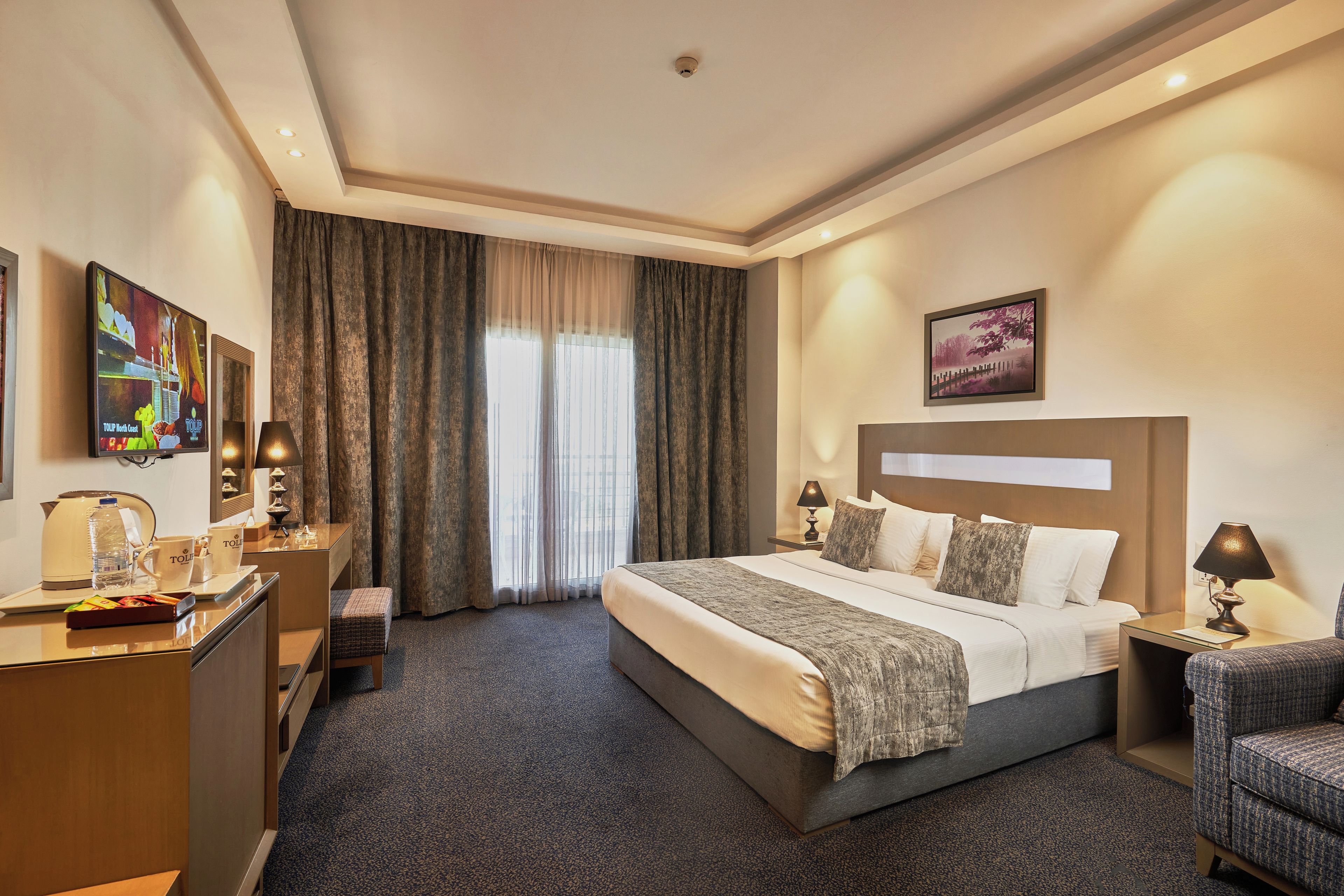 Una habitación de hotel con una cama grande, un escritorio con lámpara, una silla, un televisor y un cuadro en la pared.